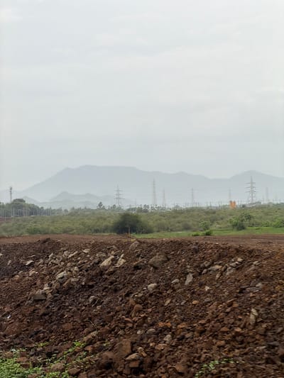 The image depicts a landscape with a foreground of dark brown soil and rocks, suggesting a construction site or a rural area undergoing development. In the mid-ground, a line of dense green vegetation runs horizontally, with trees and shrubs. Beyond the vegetation, a series of tall, metal electrical towers are visible, indicating infrastructure. In the background, a range of hazy, mist-covered mountains stretches across the horizon, with the sky appearing overcast and light gray. The scene conveys a sense of a vast, somewhat undeveloped area with a mix of natural and man-made elements. The location is specified as Vasai-Virar, India, suggesting a context within that specific urban or semi-urban region.
