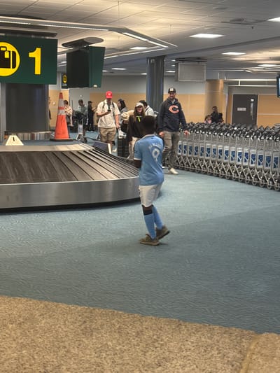 In Richmond, Canada, at an airport baggage claim area, a young boy wearing a light blue soccer jersey with the name "HARLAND" and the number "9" on the back, along with white shorts and blue socks, stands facing away from the camera. He is wearing athletic shoes. In the background, a baggage carousel is visible, along with other travelers waiting and a row of luggage carts. The floor is carpeted in a blue geometric pattern. Overhead, a sign displaying a yellow suitcase icon and the number "1" is illuminated. The lighting suggests an indoor, well-lit environment typical of an airport terminal.