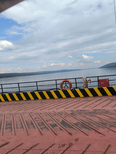 The image depicts a view from a ferry or barge on a body of water, likely the Angara River, near the town of Boguchany, Russia. The foreground is occupied by the deck of the vessel, which is painted a faded red with a textured surface of parallel grooves and raised elements. A safety railing, painted black with diagonal yellow and black hazard stripes, runs along the edge of the deck.
Beyond the railing, a wide, calm body of water stretches to the horizon. Distant, rolling hills covered in dark green vegetation form the landscape on the opposite shore. The sky is partly cloudy, with patches of blue interspersed with large, fluffy white clouds, suggesting fair weather.
Attached to the railing are several objects. A bright orange life preserver ring is visible, as is a coiled section of beige hose connected to a red fire hydrant. A red metal box, possibly a fire safety cabinet labeled "ПР-2," is also mounted on the railing. The overall impression is one of a peaceful, scenic journey on a waterway, common in the region of Boguchany.