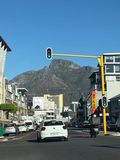 This image depicts a vibrant daytime street scene in an urban setting, with a prominent mountain in the background, likely Table Mountain in Cape Town, South Africa. The photograph is taken from a slightly elevated perspective, looking forward along a city street.

In the foreground and midground, a busy street is visible with parked cars on the right side. A white Volkswagen Polo with the license plate "CY 320-720" is closest to the viewer. Further down the street, a delivery motorcycle with a large black box on the back and a license plate ending in "435FS" is also visible. Overhead, a bright yellow pole extends across the street, supporting two traffic light units, both showing green signals, indicating that traffic can proceed.

Buildings line both sides of the street, featuring various architectural styles and multiple windows. In the midground, several signs are displayed on the buildings. One sign clearly shows "up&up group" in orange text. Another advertisement for "CAPITEC Bank" reads "Cape Town, Why pay more? Bank on better," accompanied by an image of a phone, reinforcing the location as Cape Town.

The background is dominated by the imposing, craggy slopes of a large mountain under a clear, bright blue sky, suggesting sunny weather. The overall scene captures the blend of urban life with stunning natural scenery characteristic of Cape Town.