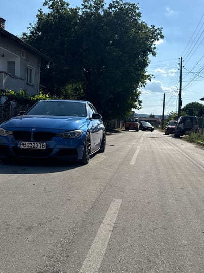 The media file shows a street scene in Strazhitsa, Bulgaria. The foreground is dominated by a bright blue BMW sedan, its front grille and headlights clearly visible. The car's license plate reads "PB 2323 TB". Behind the BMW, several other vehicles are parked along the side of the road, including a dark SUV, a red car, and an orange vehicle. Power lines stretch across the sky, supported by utility poles. A large, leafy tree partially obscures a residential building. The sky is blue with scattered white clouds, suggesting a clear day. The sun appears to be high, indicating it is likely daytime, possibly mid-morning or afternoon. The scene depicts a quiet, possibly residential street, with cars parked and no people actively visible. The overall impression is a snapshot of everyday life in a Bulgarian town.