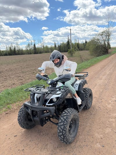 A person wearing a helmet and light-colored clothing is sitting on a black and white ATV on a dirt road next to a grassy field. The sky is blue with scattered white clouds. Trees are visible in the background. The ATV is facing towards the viewer, slightly angled to the right. The person is looking at the camera. Based on the environment, the location could be a rural area near Mozhga, Russia, given the presence of a dirt road, fields, and trees, which are common landscapes in the region.