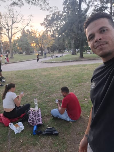 This media file is a selfie taken by a man in a public park, rotated 90 degrees clockwise. The scene takes place in Villa Domínico, Argentina, during the late afternoon or early evening, evidenced by the warm, golden light of the sun filtering through the trees in the background.
In the foreground, the man taking the selfie has short dark hair, light stubble, and wears a black t-shirt with an unreadable white logo. A tattoo is visible on his left forearm as he looks at the camera with a calm expression.
Behind him, to the left, a man in a red t-shirt and light blue jeans is seated on the grass, focusing on his smartphone. Further left, a woman with dark hair tied back, wearing a white short-sleeved top and dark pants, kneels on the grass, also appearing engaged with an item on the ground.
Personal belongings are scattered on the grass near the woman, including a red padded jacket, a white plastic bag with contents, a pink plaid tote bag with the stylized text "DOODLE" or similar, a blue water bottle, and a clear plastic bottle. A dark messenger bag lies near the selfie-taker.
The setting is a green public park featuring a paved walking path and numerous trees, some deciduous with bare branches and others with full green leaves. Several indistinct figures can be seen in the distance, walking or sitting, indicating a casual, relaxed atmosphere in the park.