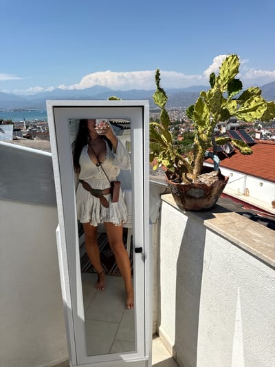 In the city of Unknown, Türkiye, a woman stands on a rooftop balcony, taking a selfie in a full-length mirror. She is wearing a light-colored, short dress, a brown belt with an ornate buckle, and a small brown pouch. The reflection in the mirror also shows her holding a pink object to her face, possibly a flower or a phone case. The background of the scene is a panoramic view of a coastal town with red-tiled roofs, mountains in the distance, and a clear blue sky with scattered clouds. A large potted cactus is positioned on the right side of the balcony, adding a touch of desert flora to the urban landscape. The balcony's white textured wall and a marble-like ledge frame the scene, suggesting a warm, sunny climate.