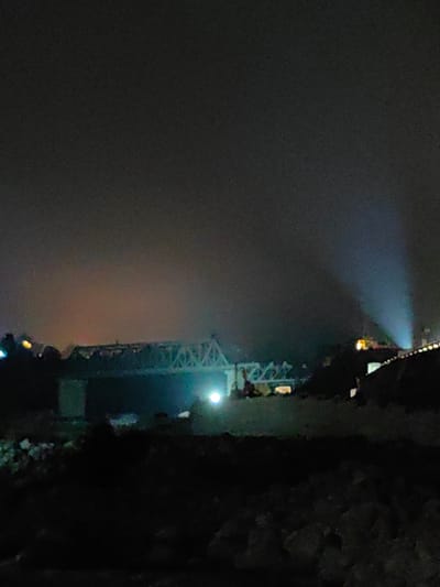 The image is a nighttime photograph of a bridge in Kulang, India. The bridge, a truss bridge, is illuminated by lights and spans a body of water. The sky is dark and cloudy, with some light pollution visible. In the background, there are some lights from buildings or other structures. The foreground is dark and appears to be a rocky or muddy area. Two beams of light are visible in the sky on the right side of the image, suggesting the presence of spotlights or searchlights. The overall impression is one of a somewhat industrial or urban landscape at night.