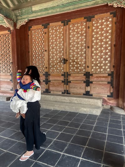 A woman is carrying a baby in a blue and white traditional Korean outfit. The baby is wearing a colorful striped sleeve top and has their mouth open and arms raised. The woman is wearing a black skirt and white flip-flops. They are standing on a tiled floor in front of large wooden doors with intricate latticework. The background features architectural elements with painted decorations. This scene is likely within the grounds of Gyeongbokgung Palace in Seoul, South Korea, given the architectural style and traditional attire.