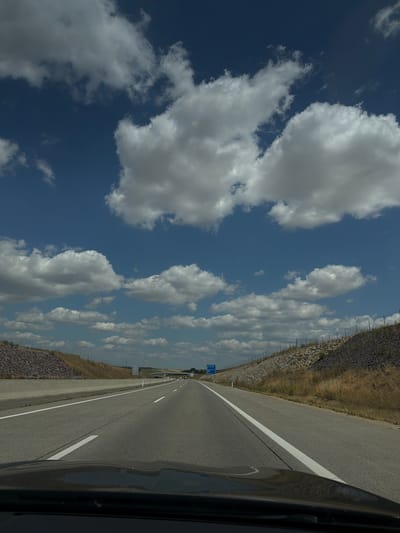 The image depicts a view from the interior of a vehicle driving on a multi-lane highway. The perspective is from the driver's seat, looking forward. The highway stretches into the distance under a partly cloudy blue sky. The road is paved with asphalt and marked with white lines. On either side of the highway, there are sloped embankments covered with dry grass and rocks. A concrete barrier separates lanes in some sections. A blue highway sign with white text is visible on the left side of the road in the distance. The hood of the vehicle is visible in the foreground, reflecting the sky. The scene appears to be in a rural or semi-rural area, given the natural vegetation and the open sky. The location is identified as Poysdorf, Austria.