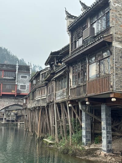 The image displays a panoramic view of the Tuojiang River in Fenghuang Ancient Town, China, under an overcast sky. The calm river, a dark green hue with slight surface ripples, occupies the lower foreground and midground. Along the right bank, a series of multi-story traditional wooden stilt houses line the waterway, supported by numerous slender wooden poles extending into the river. The structures are primarily constructed from dark timber planks and dark grey brickwork, featuring traditional tiled roofs with curved eaves. Multiple windows with intricate wooden lattice patterns are visible on the building facades, along with wooden balconies. An air conditioning unit is mounted on an upper floor of one of the central stilt houses. Towards the right, the stilt structure transitions to include robust, square grey stone pillars supporting the overhang, with two white spherical lanterns suspended beneath. In the midground-left, a reddish-brown stone arch bridge spans the river, connecting to a prominent grey building with red trim and dark roofing, also featuring traditional architectural elements. Distant forested hills, partially obscured by atmospheric haze, are visible in the background. No people are present in the scene.