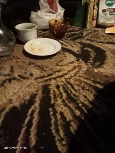 The image displays an indoor scene on September 6, 2025, at 20:08, within an unidentifiable specific location in the Cotswold District, United Kingdom. The foreground features a dark brown and beige patterned carpet, exhibiting scattered light-colored particulate matter. Visible objects include: a partially obscured clear glass bottle on the far left, a white ceramic mug, and a small white ceramic plate containing light-colored food residue. To the right of the plate, an amber-colored glass tumbler is present. The background contains a crumpled white plastic bag, a red object, a dark green object, a beige package with partial text, and a white package labeled "EKELSO KALOSI TORAJA." A small orange item is also visible on the far right background. No individuals are depicted. The scene is dimly lit by ambient artificial light.