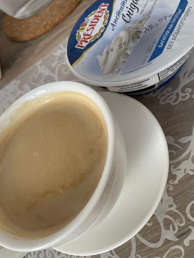 An overhead medium shot captures a coffee cup filled with frothy coffee, resting on a saucer. To the right of the coffee cup, a partially opened container of President cream cheese is visible. The container has blue and white labeling with text in Cyrillic script. The scene is set on a table with a patterned placemat. The background is blurred but suggests a domestic setting, possibly in Brovary, Ukraine. No people are visible in the image. The lighting is natural, and the overall composition is a still life of breakfast items.