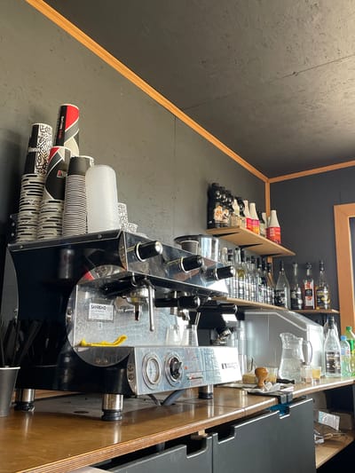 The image depicts a coffee machine and surrounding counter area in a commercial setting. The coffee machine, a black and stainless steel Sanremo model, is positioned on a wooden countertop. To the left of the machine, stacks of disposable cups are visible. Above the counter, shelves are stocked with various bottles, including syrups and alcoholic beverages, as well as jars and other items. The background consists of a dark grey wall and a dark grey ceiling with wooden trim. There are no people visible in the image. The scene appears to be a clean and organized coffee preparation area. The image does not contain any specific visual cues that would allow for identification of its location in Shiryaevo, Russia.