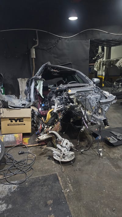 The image depicts the aftermath of a severe car accident within an indoor workshop or garage setting in Ataşehir, Turkey. The primary subject is a heavily damaged vehicle, likely a car, that has been stripped down and disassembled. The vehicle's chassis, engine, and various internal components are exposed, with numerous wires, cables, and mechanical parts strewn around it. Some parts appear to have been removed or are hanging loose, suggesting an ongoing repair or dismantling process.

In the background, the workshop environment is visible, characterized by concrete flooring, dark walls, and overhead lighting. Some stacks of debris or parts are also present, contributing to the chaotic scene. To the left of the vehicle, a bright white light source, possibly a work lamp, illuminates the area.

Among the scattered automotive parts, a cardboard box with visible text stands out. The text on the box reads "TYC LIGHTS, PRECISION SAFETY AUTOMOTIVE LAMPS". Other visible text is limited and difficult to discern.

There are no people visible in the frame. The scene conveys a sense of disarray and damage, indicative of a significant vehicular incident. The time of day cannot be determined, but the artificial lighting suggests it is likely indoors and potentially not during daylight hours. The weather is not discernible as the scene is indoors.