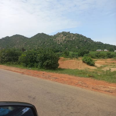 The image captures a view from a moving vehicle, looking towards a lush green, rocky hillside. In the foreground, a paved road with a reddish-brown dirt shoulder is visible, with the side mirror of the vehicle in the bottom left corner. The hillside is densely covered with vegetation, featuring a mix of trees and shrubs, interspersed with large rocks and boulders, particularly on the upper slopes. Scattered amongst the greenery are a few small buildings, appearing to be houses with red and white roofs, suggesting a small settlement at the base of the hills. The sky above is partly cloudy, with patches of blue sky and white clouds. The environment appears to be a rural or semi-rural area in Dorowa, Nigeria, characterized by its hilly terrain and vegetative cover.