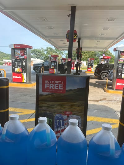 This is a medium shot, eye-level, outdoor photograph taken at a Pilot gas station in Raphine, Virginia, United States. The foreground is dominated by a row of large, clear plastic jugs filled with bright blue liquid, likely windshield washer fluid, lined up on the ground. Behind the jugs, a prominent sign displays a "BUY 2 GET 1 FREE" promotion for Monster Energy drinks, featuring images of the beverages and a landscape.
In the midground, several red and white Pilot gas pumps are visible, with some cars parked at them, including a black pickup truck and a dark-colored SUV. The pumps have digital displays and branding. Yellow lines on the asphalt indicate parking and driving lanes.
The background shows a treeline and a gently sloping hill under a bright, overcast sky. The overall scene depicts a typical gas station environment with a focus on promotional items and fueling infrastructure. There are no people clearly visible in the image.
