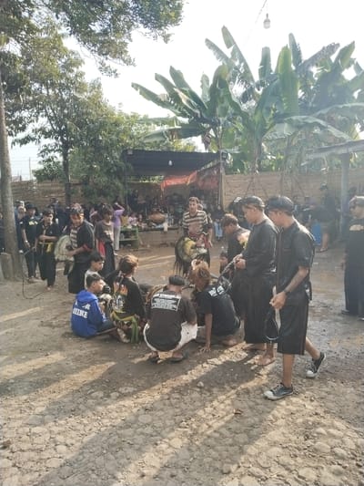 The image depicts a gathering of people in what appears to be Mojodoyong, Indonesia. The ground is uneven and dirt-covered, with some muddy patches. A group of people are clustered in the foreground, some are kneeling or squatting, while others stand or walk. The people seem to be performing some sort of ritual or activity that could be cultural or religious. Many of the people are wearing dark clothes and headbands. A crowd of onlookers is visible in the background, standing underneath a sheltered area. Banana trees and other vegetation are also present in the background.
