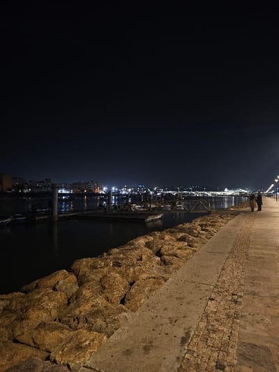 The image captures a nighttime scene along the Bou Regreg riverfront in Salé, Morocco. A paved pedestrian promenade, consisting of concrete slabs bordered by a strip of small, rectangular pavers, occupies the right side of the frame. To the left of the promenade, a retaining wall constructed from large, irregularly shaped, light brown to tan rocks forms the edge of the river. The dark water of the river contains a section of wooden floating docks or pontoons, with multiple vertical white poles visible, suggesting a marina or mooring area. Across the river, the opposite bank is heavily illuminated by numerous artificial lights from urban structures, which are reflected on the water's surface. A prominent, elongated, illuminated structure, possibly a bridge or large building complex, is visible in the distant background. Two indistinct individuals are standing on the promenade in the far right midground, facing away from the camera and towards the illuminated far bank. The scene is primarily lit by the streetlights illuminating the promenade and the distant urban lights.