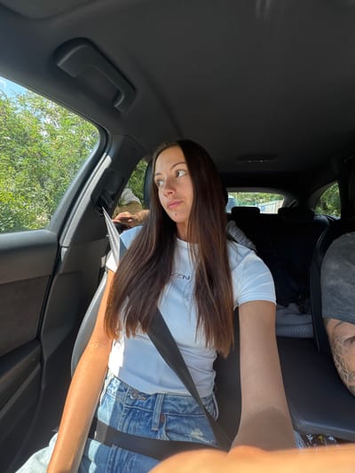 A young woman with long, brown hair is sitting in the passenger seat of a car, facing the camera. She is wearing a white t-shirt and denim shorts. The car's interior is visible, including the black upholstery, a seatbelt, and a grab handle. Through the open window, trees and a blue sky with some clouds can be seen, indicating it's daytime and likely fair weather. The woman is looking towards the camera with a slightly surprised expression. Sunlight is casting shadows on her arm and the car's interior. The scene appears to be a casual car ride, possibly during a road trip, given the outdoor scenery visible through the window. The location is Kresna, Bulgaria. There is visible text on the woman's t-shirt, appearing to be a logo or brand name, but it is partially obscured and difficult to read clearly.