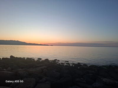The image depicts a sunrise or sunset scene at the coast in the Heraklion Municipal Unit, Greece. A body of water, presumably the sea, stretches towards the horizon. In the foreground, there are dark, rocky formations of varying sizes. The sky transitions from a pale blue at the top to orange and yellow hues near the horizon. In the distance, a mountain range is visible. There is a very faint vapor trail of an airplane is visible in the sky. At the bottom left corner of the image, the text "Galaxy A55 5G" is superimposed.