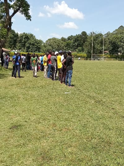 The image captures an outdoor scene on a sports field in Kakamega, Kenya. Approximately 30 to 40 individuals, predominantly African males, are gathered in a cluster on the left side of the frame, standing on a green grass surface at the edge of the field. They are dressed in various casual clothing, including t-shirts, shirts, trousers, and jeans, with some wearing headwear. The group faces generally towards the right, appearing to observe or await an event on the field. The foreground consists of a mown green grass field. In the mid-ground, a set of tall white H-shaped rugby goalposts and a shorter set of football goalposts are visible. A dense treeline forms the background, with a large, mature tree featuring a prominent trunk situated on the far left. A solitary, indistinct white figure is visible in the distant center of the field, near the goalposts. The sky above is blue with scattered white cumulus clouds, and the scene is brightly illuminated by natural sunlight, indicating daytime.
