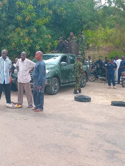 A group of men, some in civilian clothes and some in military uniforms, stand on a paved road in front of a truck. The truck is dark green and has a flatbed. There are two flat tires on the ground, one next to the truck and one further away. Behind the group of men are trees and dense vegetation. The truck is parked facing the right side of the scene. A motorbike is visible behind the men in military uniform. The men appear to be in conversation. The scene is in Ungwan Tanko, Nigeria at latitude: 10.422, longitude: 7.49.