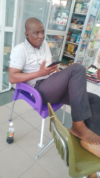 A Black man is sitting in a plastic purple chair, looking at a tablet device he is holding in his right hand. He is wearing a light gray polo shirt with a subtle pattern and dark gray pants. He has earbuds in, with the wire visible. The man has a bald head and appears to be middle-aged, with a neutral to slightly concerned expression. His left foot is resting on the seat of a green chair to his right.
To the left of the purple chair, on the tiled floor, is a plastic bottle of Coca-Cola. Behind the man, a tall display case filled with various products is visible. These products appear to be snacks, drinks, and possibly some health or personal care items, suggesting the setting is a small shop or convenience store. The lighting suggests it is daytime. The environment is indoors, as indicated by the display case and the tiled floor. The overall scene appears to be a casual moment of rest or engagement with technology within a commercial setting in Orerokpe, Nigeria. The items in the display case are too small to be clearly identified, but brand names such as "RAMBO" and "24PKTS" are partially legible on some packaging.