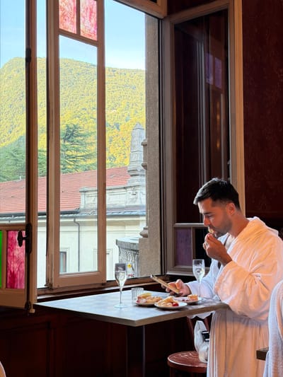 A man in a white bathrobe is seated at a table in what appears to be a hotel or spa lobby in San Pellegrino Terme, Italy. He is looking down at his phone and eating. On the table in front of him are two glasses of champagne, a plate with various small pastries, cheese, and some fruit. The table is set against a backdrop of a large window that overlooks a lush, green mountainside with some buildings visible. The window has stained glass inserts in the upper section and a dark wood frame. The interior of the room has dark wood paneling.