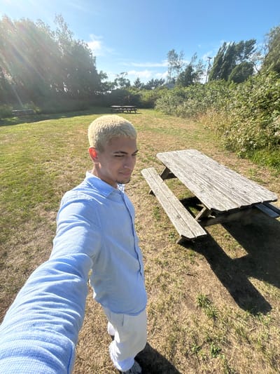 A light-skinned man with short blonde hair stands on a grassy field, taking a selfie. He's wearing a light blue button-down shirt and white pants. Behind him is a wooden picnic table casting a shadow on the ground. Other picnic tables can be seen further in the background near the trees. The sky is blue with scattered clouds, indicating a sunny day. The trees and greenery surrounding the field create a natural backdrop.