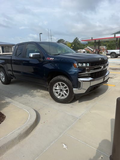 A dark blue Chevrolet Silverado pickup truck is parked in a concrete lot. The truck is facing towards the right side of the frame. The truck has chrome accents on the grille, bumper, and side mirrors. The Z71 badging is visible on the front fender. In the background, a gas station with a red and white canopy is visible, along with some trees and buildings. The sky is cloudy. The scene is consistent with a gas station parking lot in Chattanooga, Tennessee, United States of America.