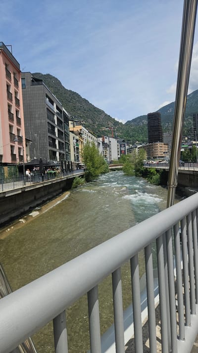 The image presents a detailed view of the Gran Valira river flowing through Andorra la Vella, Andorra, observed from a bridge on a clear, sunny day. In the immediate foreground, a grey metal bridge railing, composed of a horizontal top bar and numerous vertical balusters, extends from the lower right across the bottom of the frame. To the railing's left, a reflective silver-colored vertical support column is partially visible.
The midground is dominated by the river, characterized by its murky, brownish-green water and numerous areas of turbulent white rapids, indicative of a strong current. The left bank is densely urbanized, featuring a promenade lined with buildings. The closest structure on the left is a multi-story light pink building with balconies. Adjacent to it stands a modern, dark grey building with extensive fenestration and balconies. Further along the bank, a series of diverse commercial and residential buildings of varying colors and styles are visible. Below these structures, an outdoor cafe area with multiple black umbrellas and seated patrons is active, and several individuals are seen walking along the promenade.
The right bank, extending into the distance, also features urban development, including a tall, dark brown building prominent against the mountainous backdrop. Green foliage and trees line sections of both river banks. Two bridges span the river in the distance: a smaller, light-colored bridge closer to the center, and a wider bridge further downstream. A red construction crane is discernible high on the mountain slope on the right side of the image. The background consists of steep, verdant mountains, covered in trees and exposed rock, rising directly behind the city. The sky above is largely clear and blue, with scattered white clouds.