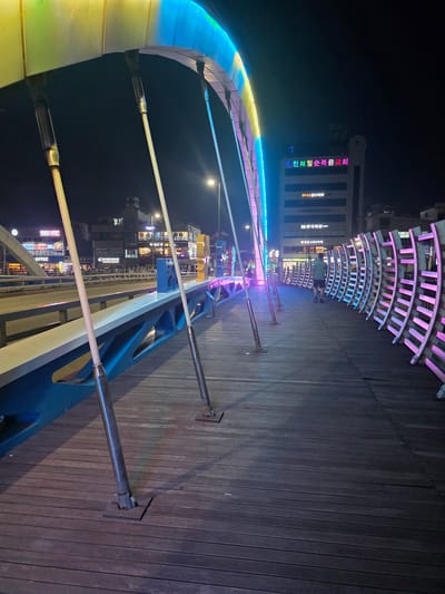 The image depicts a pedestrian bridge at night in Eumnae-ri, South Korea. The bridge features a colorful, illuminated archway that casts vibrant light on its wooden deck. Several slender poles, likely structural supports for the arch, are visible along the walkway. The walkway itself is constructed of dark wooden planks, with metal bases securing the poles to the surface. In the background, buildings with illuminated signage are visible, including what appears to be a church with Korean text and a cross emblem. A person, seen from behind, is walking away from the camera on the right side of the bridge. The overall environment is urban and nocturnal, characterized by artificial lighting.