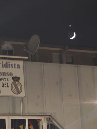 An image captured from an elevated perspective looks towards a building's facade at night, with a clear view of the sky above. In the sky, a crescent moon is visible, along with a bright point of light, likely a planet or star. Below the sky, a satellite dish is mounted on the building. A sign with purple text is affixed to the facade, partially obscured by the angle. The text on the sign reads "ridista," "onso," "NTE," and "DEL." The Real Madrid club crest is also present on the sign. Below the sign, a windowed area is visible, with colorful triangular flags hanging inside, suggesting a festive atmosphere or decoration. The overall scene depicts an urban nightscape with celestial observation and building elements. This image was taken in Cornellà de Llobregat, Spain.