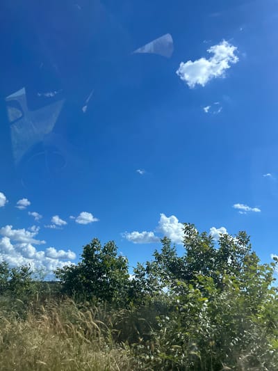 The image shows a bright blue sky with scattered white cumulus clouds. In the foreground, there are lush green trees and tall, dry grass. The scene appears to be a natural landscape, likely in a rural area, and there is a reflection on the left side of the image, suggesting it was taken through a window. There are no people or specific landmarks visible that would indicate it is in Konstantinovo, Bulgaria.
