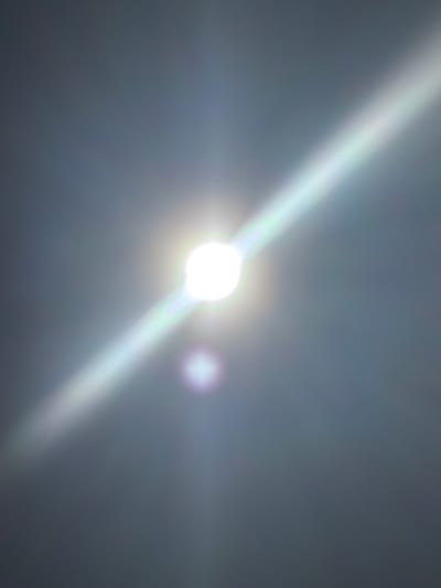 The image is an abstract, blurry depiction of a bright light source, likely the sun, creating lens flares that extend diagonally across a dark, muted background. There are no discernible objects, people, or environmental features that would suggest a specific location within the city of Karachino, Russia, or any other identifiable setting. The light source is a central, bright white circle with rays of light radiating outwards, some appearing as white streaks and others exhibiting faint rainbow-like colorations, particularly green and purple. There is a secondary, smaller, and more diffuse light flare present in the lower portion of the image, also with hints of purple. The overall impression is one of atmospheric phenomena rather than a concrete scene.