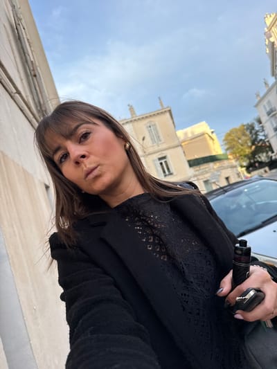 A woman in a black lace top and sweater holds keys and a vape pen. Buildings stand tall in the background against a blue sky, with glimpses of green foliage. The image has been rotated, tilting the woman's head to the side. The angle offers a low perspective, looking up at the buildings and sky.