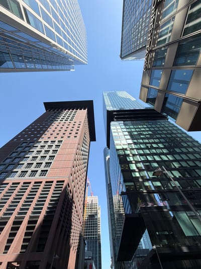 This is an outdoor, eye-level, wide-angle photograph taken from below, looking up at a cluster of modern skyscrapers against a clear blue sky. The perspective creates a sense of immense scale and architectural grandeur. The buildings are a mix of glass and stone, with clean lines and geometric patterns. Some buildings have reflective glass facades that mirror the sky and surrounding structures, while others are clad in reddish-brown stone with rows of windows. The time of day appears to be daytime, with bright sunlight casting distinct shadows on the buildings, highlighting their textured surfaces. There are no people visible, and the focus is solely on the imposing architecture.
One of the prominent buildings, a tall, reddish-brown structure, has a distinctive stepped silhouette and a grid-like pattern of small windows or vents. Another, a lighter-colored glass skyscraper, shows a repeating pattern of large, rectangular windows. In the background, partially obscured by the foreground buildings, are other structures, including one under construction with visible cranes.
There are a few instances of visible text: "MAYER.BRÖ." is partially visible on a reflective surface of one building, suggesting a company name. Another building's facade displays the word "SPACES." in white lettering. The overall impression is one of a modern, bustling urban environment, characteristic of a major financial district. The clear sky and strong sunlight suggest good weather. The upward angle emphasizes the height and impressive scale of the architecture.