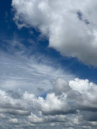 The image displays a dynamic sky filled with a variety of clouds against a deep blue background. In the upper left, a large mass of white, fluffy clouds dominates the frame, with its edges softly blending into the blue. Below this, a layer of wispy, thin clouds stretches across the middle of the image, creating a feathery texture. Further down, more substantial, textured clouds are visible, some appearing dark and heavy, suggesting potential for rain, while others are bright white. The lower portion of the image is densely packed with these varied cloud formations, creating a sense of depth and volume. The scene is a natural sky, indicative of weather conditions possibly experienced in or around Ol'khovskoye, Russia. There are no people, animals, or man-made structures present in the image.