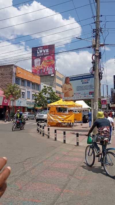 A busy street scene features a KFC billboard advertising "LOVE AT FIRST BITE," and a "BLOOM MAMA" advertisement for comprehensive maternal care. A yellow "Sun King" tent promotes their new EZI smartphone. People are visible walking and riding bikes, including one cyclist in the foreground. Buildings with signage such as "JUMLADJUMADRA," "Itel," and "TAI SACCO" line the street, with utility poles and wires overhead against a partly cloudy sky.