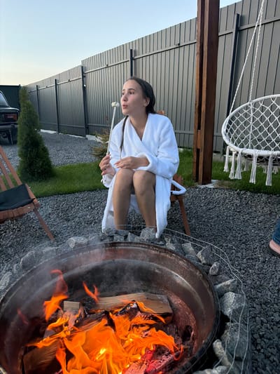 In Sharovechka, Ukraine, a young woman wearing a white bathrobe is seated in a wooden chair in an outdoor area. She is holding a metal skewer with a white object on it, and her mouth is open as if she is about to eat or drink. A fire burns in a metal fire pit in the foreground, surrounded by rocks. Behind the woman, a dark metal fence encloses the area, and a wooden post supports a white hammock chair. A dark-colored vehicle is partially visible to the left, and a small evergreen tree stands near the fence. The ground is covered in dark gravel and patches of grass. The sky is a pale blue, indicating it is either early morning or late evening.