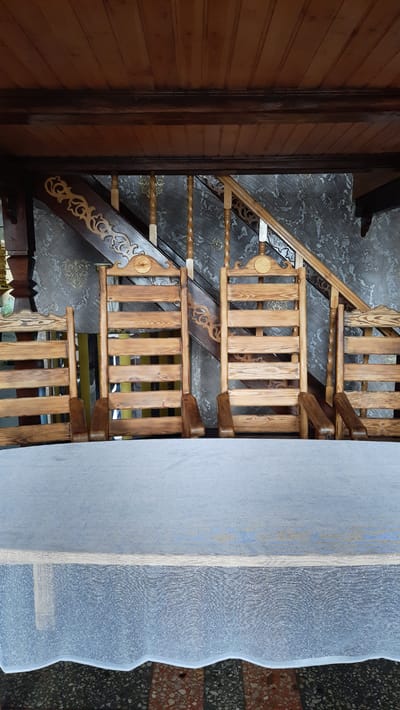 In a covered outdoor seating area, possibly a veranda or pavilion, the scene is dominated by wooden furniture and architectural elements. Four light-brown wooden chairs with slatted backs and armrests are arranged in a row. The chairs are positioned behind a round table draped with a light-blue, textured tablecloth that has a white, patterned border along the bottom. Above the chairs, a wooden ceiling with visible planks and beams forms the roof. A wooden staircase with intricately carved wooden balusters and railings ascends in the background, with a patterned wallpaper on the wall behind it. The flooring visible beneath the table appears to be a mosaic of colored stones. The overall impression is of a rustic, possibly traditional, outdoor dining or seating area. The location is identified as Жодзішкаўскі сельскі Савет, Belarus.
