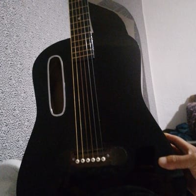 The image shows a close-up of a black acoustic guitar positioned in front of a textured, light-colored wall. The guitar has six strings that run vertically down its body. The soundhole is oval-shaped and has a silver trim. The bridge of the guitar, with seven white bridge pins, is visible towards the bottom of the frame. A portion of a person's hand, with a thumb and index finger visible, is resting on the lower bout of the guitar, suggesting someone is holding or playing it. The background wall appears to be a patterned wallpaper. The lighting suggests an indoor setting. There are no discernible landmarks or clues to indicate the location within the city of Vasylkiv, Ukraine.