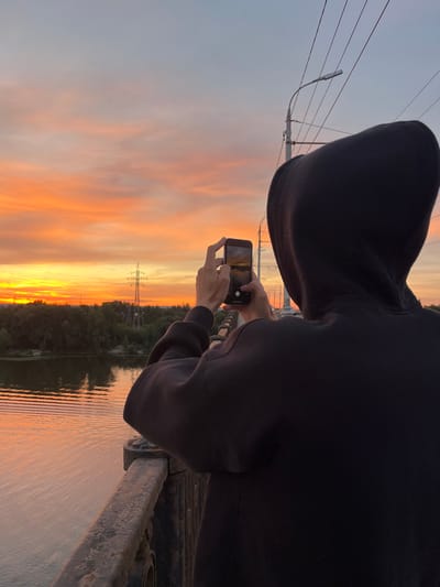 A person in a black hooded sweatshirt is holding up a smartphone to capture a photo of a sunset. The phone is oriented vertically, and the person's hands are visible as they operate the device. The sunset is vibrant, with hues of orange, yellow, and pink painting the sky. A body of water, likely a river, reflects the colors of the sunset. Trees and a power line tower are silhouetted against the colorful sky on the far bank of the river. The person appears to be standing on a bridge or elevated platform, as indicated by the metal railing on the right side of the image. The time of day is clearly sunset, and the weather appears clear. The overall mood is serene and appreciative of the natural beauty. No visible text is present in the image. The location is identified as Samara, Russia.