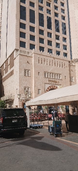 The image displays the ground-level entrance of Tower 3, part of a multi-story building complex in Makkah Al Mukarramah, Saudi Arabia. The building, constructed from light-colored stone or facade material, features multiple rectangular windows and ornate lower sections with decorative lattice-patterned windows and crenellated rooflines. The designation "TOWER 3" is prominently inscribed above a large arched entrance. In the foreground, a black SUV is parked on a patterned paved surface to the left. A light-colored canvas canopy extends over an outdoor area on the right side of the frame. Beneath and adjacent to the canopy, several metal carts are present, including a blue utility cart carrying items and a traditional bellhop-style luggage cart. Two individuals are partially visible: one near the blue utility cart and another further back under the canopy. A decorative street lamp with multiple lanterns stands on the left. Shadows indicate daylight conditions.
