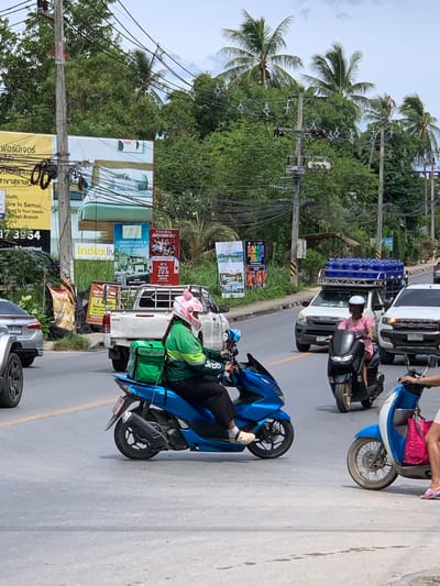 The image depicts a street scene in Baan Bo Phut, Thailand, with several vehicles and pedestrians. The dominant elements are motorbikes, a white pickup truck, and a blue delivery vehicle. Two individuals are visible on motorbikes. One person, wearing a pink shirt, is riding a black motorbike. Another person, in a green jacket and helmet, is on a blue motorbike, appearing to be a delivery driver with a green bag strapped to the back. The blue delivery vehicle has "Grab" branding visible on its side.
The setting appears to be a roadside with palm trees and dense greenery lining the street. Several billboards and advertisements are posted along the road, some in Thai script, indicating a commercial or populated area. One billboard advertises "LUXURY VILLAS & APARTMENTS FOR RENT" and another mentions a "GRAND FRAND SALE UP TO 70% KICK OFF DEALS." There are also utility poles with numerous wires overhead.
The time of day seems to be daytime, with bright lighting suggesting clear or partly cloudy weather. The overall atmosphere suggests a busy, everyday scene in a tropical location. The road surface appears to be asphalt, with a yellow line indicating a lane divider. The image is oriented vertically, with the bottom of the scene appearing at the top of the frame, suggesting it might have been rotated.