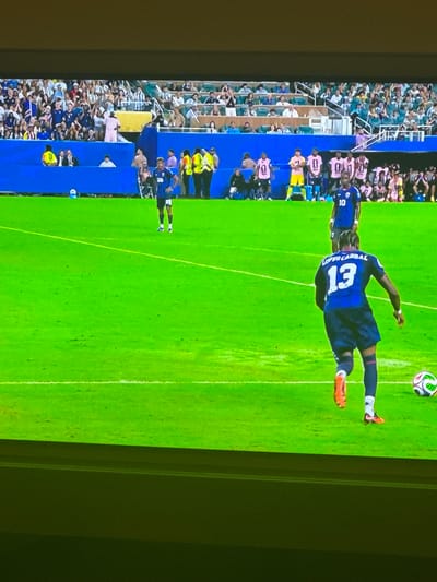 The image depicts a soccer match on a bright, sunny day. The playing field is a vibrant green, with white lines marking the boundaries. In the foreground, a soccer player wearing a blue jersey with the number 13 and the name "LOPEZ CARRIBAR" on the back is positioned near a soccer ball. He is in the process of kicking the ball, with his right leg extended and his left leg planted on the ground. His jersey has a white trim around the neckline and sleeves, and he is wearing dark shorts and dark socks with white accents. The player's cleats are orange and white. In the background, other players in blue and pink jerseys can be seen, along with spectators in the stands. The stadium appears to be modern, with tiered seating. There is no visual information in the image that would indicate a location in Sarıyer, Turkey.