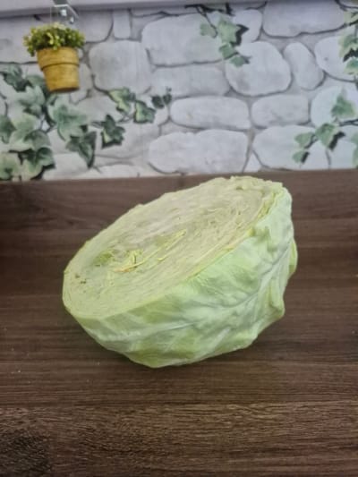 This image shows a close-up, overhead shot of a halved head of iceberg lettuce placed on a dark wood surface. The lettuce is fresh, with tightly packed pale green leaves visible in the cross-section. The outer leaves are a lighter, more vibrant green. To the left of the lettuce is a backdrop with a printed pattern of light gray stones and green ivy leaves. In the bottom left corner of the frame, a small, yellow decorative pot filled with artificial green foliage is visible, attached to what appears to be a white surface. The lighting suggests an indoor setting, possibly a kitchen counter, with no specific time of day or weather cues. There is no visible text in the image. The overall impression is a simple, clean presentation of a fresh vegetable.