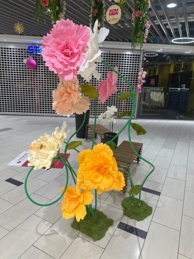 This media file showcases a vibrant, large-scale paper flower installation in what appears to be an indoor public space, possibly a shopping mall or atrium. The scene is brightly lit, suggesting daytime.

The primary subjects are an assortment of oversized, colorful paper flowers in shades of pink, peach, yellow, and cream. These flowers are arranged on a green wireframe structure that mimics stems and branches, creating a whimsical and artistic display. Lush green moss-like decorations are also integrated into the installation, adding texture and a natural element.

The background reveals a modern tiled floor and a store facade with glass windows, reflecting the surroundings and overhead lighting. There are hints of a retail environment with display shelves visible through the glass. In the distance, a wooden bench offers seating, suggesting a place for people to rest and admire the display.

One prominent piece of text is a round yellow tag with red lettering that reads "SOM MER morenen". There is also a red and white sign with text that appears to be in a Scandinavian language. Another sign is partially visible with blue lettering, "STS". Small pink balloons are also suspended in the background.

The overall impression is one of cheerful decoration, likely for a seasonal event or to enhance the aesthetic appeal of the location. There are no people visible in the image, focusing the attention entirely on the artistic floral arrangement.