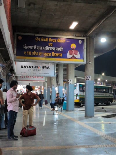 The image captures a scene at a bus station, likely in Kajheri, India, based on the signage. In the foreground, two men are engaged in conversation. The man on the left wears a light pink shirt and dark blue jeans, with a black turban. The man on the right has a maroon-colored shirt, light beige pants, and a black turban. A red duffel bag rests on the floor between them.

Behind them, a large blue banner advertises "RAYAT-BAHRA UNIVERSITY" with contact information and campus locations. Further back, a green and white bus is parked, with the Punjab Road Transport Corporation logo visible. Other individuals are seen scattered around the station, some waiting and others moving. The environment appears to be an open-air covered area with concrete pillars and a tiled floor. The lighting suggests it is either late evening or early morning. The overall atmosphere is that of a busy public transportation hub.