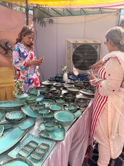 The image shows two women standing at a table displaying a variety of ceramic dishes and bowls at a market in Sector 17, Chandigarh, India. One woman, wearing a white dress with floral patterns, holds a phone in her left hand and appears to be looking at it. The other woman, wearing a white dress with red stripes, points her right hand at the table.  The table is covered with a white cloth and lined with an array of teal-colored ceramic dishes and bowls with dark streaks.  A large industrial fan is visible in the background. The setting is outdoors with a canopy overhead.
