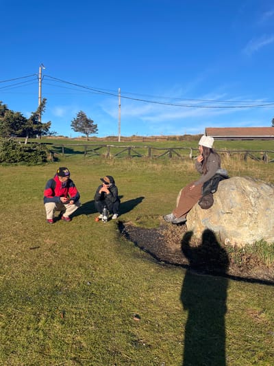 The image depicts a group of three people outdoors in a grassy area in Ushuaia, Argentina. A woman wearing a patterned beanie, brown jacket, and brown pants is standing next to a large rock. Two other people are seated on the grass: one in a red and blue jacket with a yellow hat, and another in a black jacket and hat. There is a wooden fence and a building with a brown roof in the background. The sky is blue and clear. The scene suggests a park or rural setting.