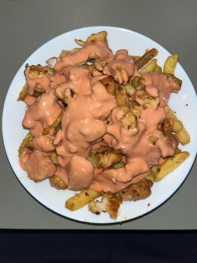 The image displays a meal on a white, round plate, resting on a light grey surface. The plate is largely filled with golden-brown, irregular-cut French fries, interspersed with numerous pieces of cooked chicken. Both the fries and chicken are heavily coated in a thick, peach-colored, viscous sauce, which has a smooth texture with minor discernible lumps and specks, possibly chili flakes. The sauce covers most of the food, obscuring some details of the chicken and fries beneath. The immediate background is a plain light grey, with a darker, indistinct horizontal band visible at the bottom edge of the frame. There are no people, explicit actions, or interactive elements visible. The image contains no visual information to corroborate the specified location of Stevenage, United Kingdom.